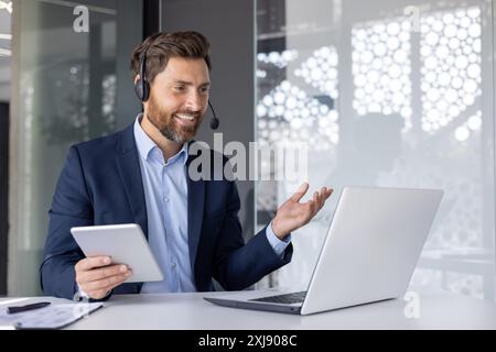 Un uomo d'affari professionista sta partecipando a una riunione virtuale utilizzando un auricolare e un computer portatile mentre tiene in mano un tablet in un ambiente d'ufficio moderno. Foto Stock