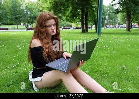 Bella ragazza con lunghi capelli di zenzero seduta con un computer portatile su un'erba nel parco estivo che lavora all'aperto Foto Stock