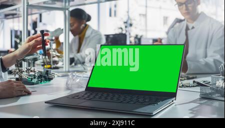 Computer portatile con schermo verde in piedi su un tavolo in un laboratorio di classe scientifica. Diversi studenti che praticano la saldatura di componenti elettronici per i loro progetti tecnologici Foto Stock