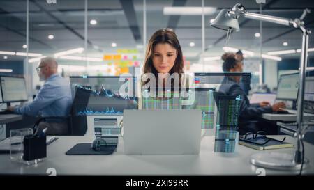 Giovane donna d'affari felice che utilizza un computer portatile in ufficio moderno con i colleghi. Elegante e bellissimo consulente finanziario che lavora. VFX Hologram Edit Visualizing Stock Exchange Interface, Opened Charts (Modifica ologramma VFX). Foto Stock