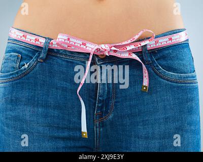 Stomaco, perdita di peso e donna con nastro, progressi e obiettivi di salute sullo sfondo dello studio. Pancia, persona e modello in jeans, dieta e benessere con Foto Stock