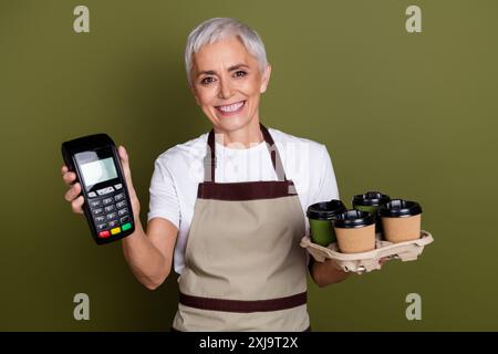 Foto di adorabile donna felice indossa un grembiule che serve bevande tenendo il terminale spazio vuoto sfondo colorato kaki isolato Foto Stock