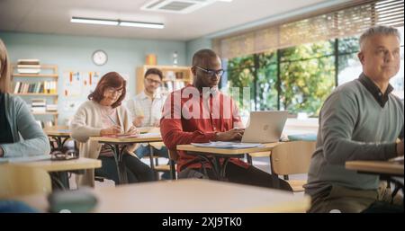 Centro di istruzione per adulti: Studenti maturi diversi che imparano in classe, utilizzano i computer portatili e scrivono in notebook. Gruppo di persone che partecipano a un workshop sul miglioramento delle competenze professionali Foto Stock