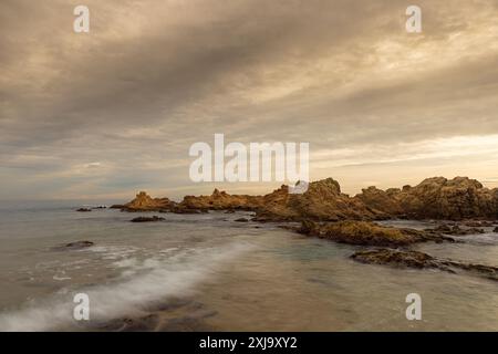 Mare di un tramonto con onde in movimento, tipiche rocce spaccate della Costa Brava, Spagna e cielo nuvoloso. Effetto del movimento sulle onde. Foto Stock