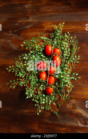 Natura morta del ramo allungato di pomodori rossi tra piccole foglie verdi su un tavolo di legno marrone. Verticale. Foto Stock