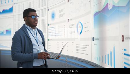Uomo d'affari di colore fiducioso che tiene in mano un notebook e che esamina i report finanziari sul grande schermo digitale nell'ufficio di monitoraggio. Imprenditore maschile che analizza la crescita e i margini di profitto di Tech Start Up Company. Foto Stock