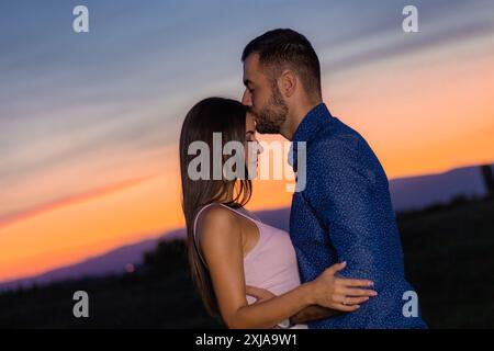 Un uomo con una camicia blu bacia una donna sulla fronte mentre stanno fuori al tramonto. Foto Stock