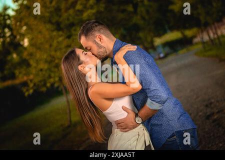 Una giovane coppia sta baciando in un parco durante il tramonto, con alberi sullo sfondo. Foto Stock