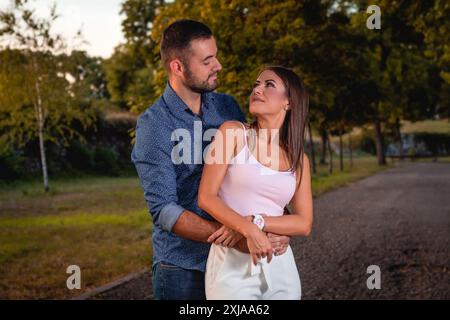 Un uomo con la barba abbraccia una donna con lunghi capelli castani. Si guardano mentre si trovano in un parco con alberi e un sentiero. Foto Stock