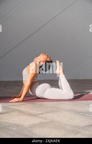 Una donna bruna vestita di abbigliamento sportivo che si esibisce in posa da yoga con il cobra reale o raja bhujangasana con luce solare che la illumina in un ambiente moderno. Foto Stock