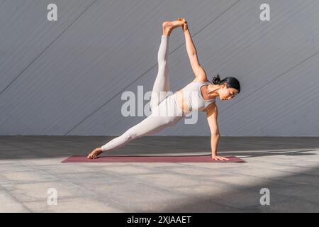 Donna bruna vestita di abbigliamento sportivo che indossa la Vasisthasana o la posa di yoga con tavola laterale. Tiene la gamba con la mano mentre fa una tavola laterale. Foto Stock