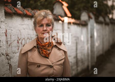 Una donna che indossa un trench marrone e una sciarpa a motivi geometrici si erge davanti a una parete bianca. Foto Stock