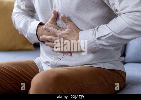 Foto ravvicinata di una parte del corpo, mani di uomini anziani che tengono il cibo, uomo in pensione che sente un forte dolore e nausea seduto sul divano di casa. Foto Stock