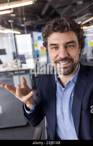 Uomo d'affari in un ufficio moderno impegnato in videochiamate guardando la videocamera sorridendo, discutendo o presentando. Ambiente professionale con arredi contemporanei Foto Stock