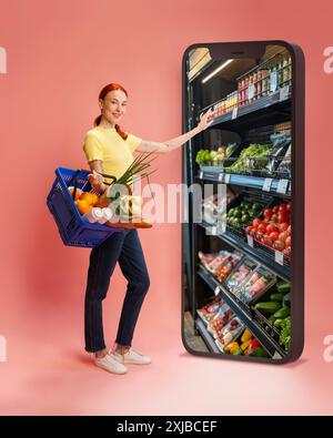 Donna con una camicia gialla che tiene in mano un cestino di alimentari, che raggiunge il gigantesco schermo dello smartphone che mostra gli scaffali del cibo su sfondo rosa Foto Stock
