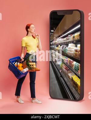 Donna in abiti casual, con cesto pieno di generi alimentari, che arriva sullo schermo gigante del telefono con scaffali della spesa su sfondo rosa Foto Stock