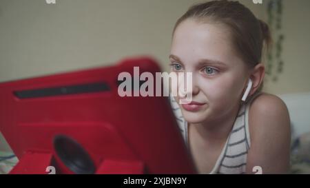 La bella adolescente con le cuffie si trova sul letto della sua stanza e naviga su Internet utilizzando un tablet digitale. Una ragazza chatta con gli amici o guarda contenuti video. Trascorrete del tempo libero in una comoda e accogliente casa. Foto Stock