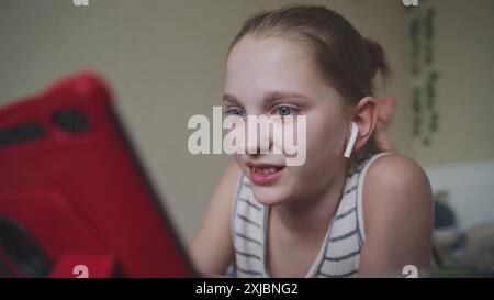 La bella adolescente con le cuffie si trova sul letto della sua stanza e naviga su Internet utilizzando un tablet digitale. Una ragazza chatta con gli amici o guarda contenuti video. Trascorrete del tempo libero in una comoda e accogliente casa. Foto Stock