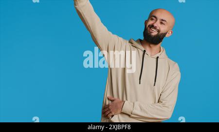 Uomo arabo che saluta una persona mentre posa in studio, si sente felice e saluta la gente su sfondo blu. Un adulto medio-orientale che si comporta spensierato e sorride con la macchina fotografica, saluta. Telecamera 1. Foto Stock