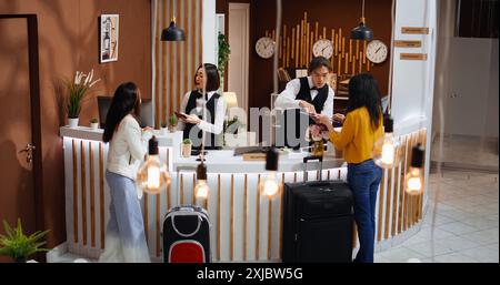 Il personale della reception aiuta gli ospiti con una facile registrazione, chiedendo di vedere le informazioni sul passaporto e di firmare i documenti di check-in per la conferma della prenotazione. Due donne in vacanza sono accolte da un personale cordiale. Foto Stock