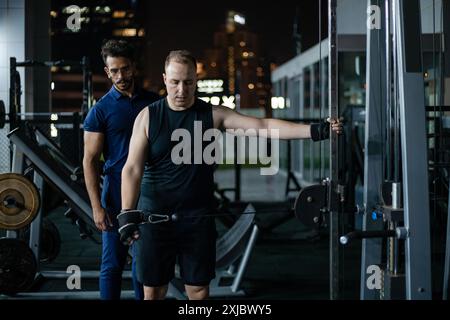 Uomo che solleva pesi con allenatore nella moderna palestra di notte Foto Stock