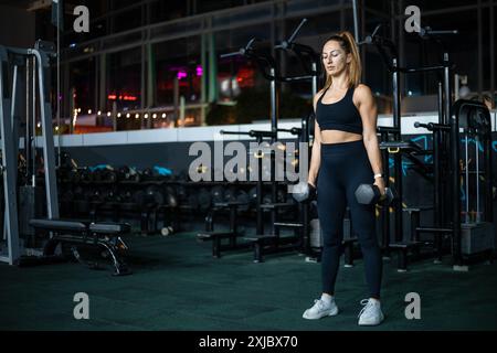 Una donna con un abbigliamento atletico nero solleva i manubri in una palestra moderna. Foto Stock