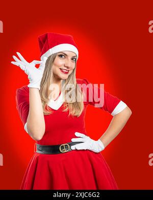 Il giovane babbo natale sorridente guarda la telecamera che fa un buon gesto di approvazione isolato su sfondo rosso Foto Stock