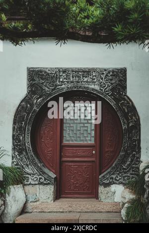 Una porta cinese tradizionale con una struttura in pietra ornata e una porta di legno rossa. La porta presenta una finestra geometrica e intricati intagli. Foto Stock
