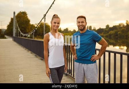 Bell'uomo e donna in abbigliamento sportivo in piedi su un ponte al tramonto e sorridente, primo piano Foto Stock