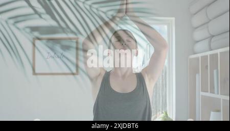 Immagine di foglie di palma verdi sopra la donna caucasica che pratica meditazione yoga in una stanza soleggiata Foto Stock