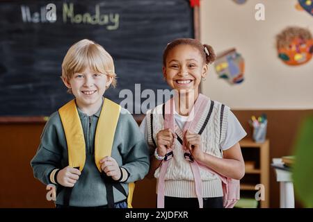 Ripresa completa media di due studenti e studentesse con zaini che sorridono e guardano la macchina fotografica in classe Foto Stock