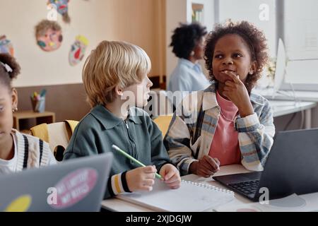 Compagni di classe multietnici che lavorano in coppia con entusiasmo mentre utilizzano un laptop seduto alla scrivania a scuola Foto Stock