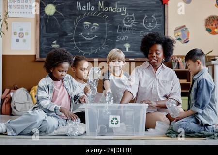 Gruppo multietnico di bambini e insegnante di sesso femminile nel club di ecologia che seleziona i rifiuti mettendo bottiglie di plastica in un contenitore di riciclaggio in classe Foto Stock