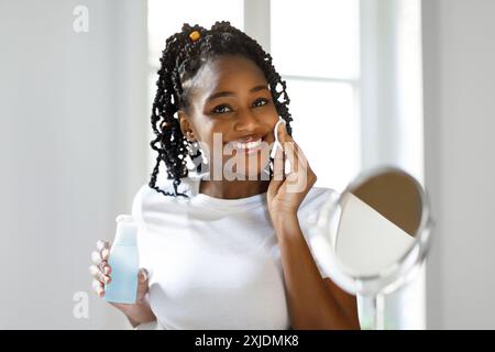 Giovane donna con trecce che applica toner alla sua faccia Foto Stock