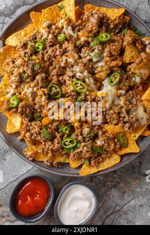 Tortilla di mais nacho messicano con carne, formaggio cheddar e pepe jalapeno serviti con salse in primo piano sul piatto. Verticale superiore vi Foto Stock