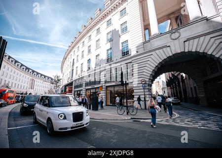 Londra, Regno Unito - 10 ottobre 2023: Un taxi bianco di Londra passa davanti a un grande edificio bianco con ingressi ad arco su Regent Street. La gente cammina sulla Foto Stock