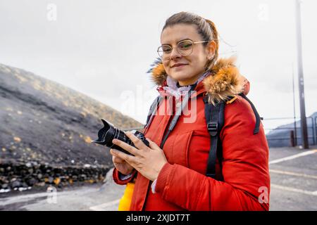 Giovane donna con fotocamera che indossa occhiali e giacca parka rossa su terreni vulcanici. Ritratto di una donna viaggiatrice e fotografa in aspre terre all'aperto Foto Stock