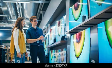 Ritratto di una donna che parla con un associato del negozio di elettronica al dettaglio. Una donna elegante cerca un televisore a schermo piatto di alta qualità. Acquirenti che esplorano le più recenti opzioni televisive nel moderno punto vendita al dettaglio Foto Stock