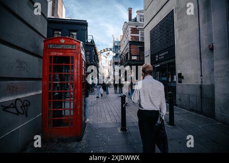 Londra, Regno Unito - 10 ottobre 2023: Una cabina telefonica rossa si trova su una strada acciottolata a Londra, Regno Unito, accanto a un uomo che cammina verso Regent Street. Foto Stock