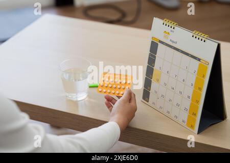 Donna che tiene in mano il calendario delle pillole anticoncezionali e un bicchiere d'acqua su un tavolo di legno Foto Stock