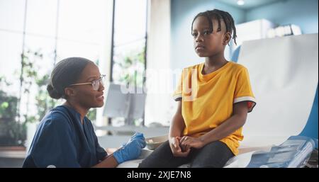 Ragazzo africano seduto sulla sedia in ospedale, parlando con infermiera nera femminile. Donna professionale che ascolta i sintomi di Young Man e dà consigli su un trattamento adeguato. Concetto di sanità pubblica. Foto Stock