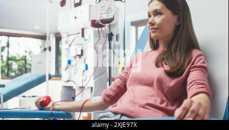 Donna caucasica donando sangue per le persone bisognose in ospedale. Donatrice che stringe la palla a forma di cuore per pompare il sangue attraverso il tubo nel sacchetto. Donazione per donne con complicazioni durante la gravidanza. Foto Stock