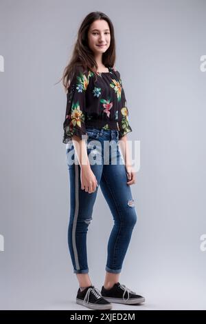 Una giovane donna elegante indossa con sicurezza jeans casual e una blusa floreale su un elegante sfondo grigio, che irradia bellezza e positività Foto Stock