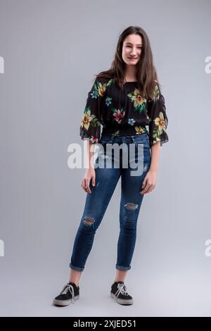 La giovane donna elegante indossa con sicurezza abiti alla moda su sfondo grigio, irradiando felicità e sicurezza con un sorriso luminoso Foto Stock