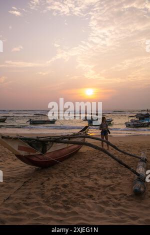 Una giovane donna gode del tramonto sulla riva dell'oceano e guarda le barche sull'acqua Foto Stock