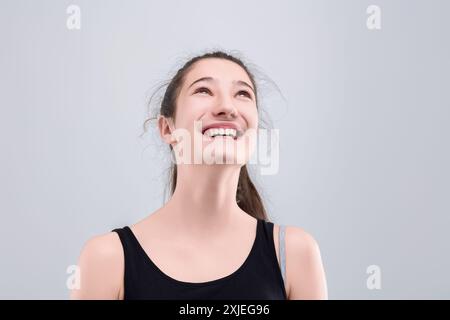 La giovane donna sorride felicemente, guardando in alto con gioia e ottimismo su uno sfondo grigio. Trasuda fiducia e speranza, perfetta per trasmettere felicita' Foto Stock