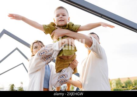 Un bambino viene trattenuto in aria dai suoi genitori. Sta sorridendo e guardando la telecamera. Le sue braccia sono allungate come se stesse volando. La sua par Foto Stock