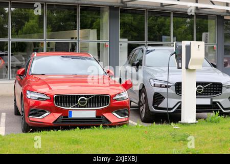 Auto elettrica ibrida rossa Volvo V60 (II) AWD 2024 collegata al caricabatteria accanto a Volvo XC60 PHEV presso la concessionaria. Salo, Finlandia. 14 luglio 2024. Foto Stock