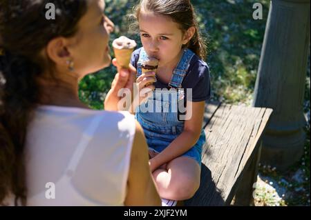 Una madre e sua figlia si godono i coni del gelato mentre si siedono su una panchina nel parco durante una giornata di sole. Il bambino indossa una tuta in denim. Foto Stock