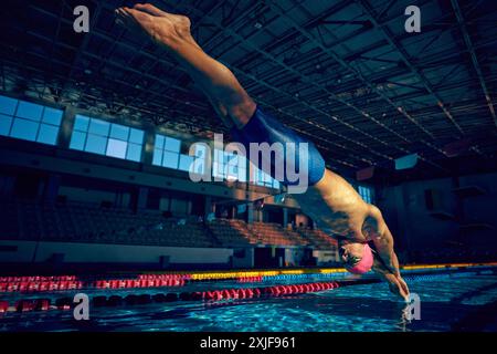 La foto dinamica dell'atleta che indossa un berretto rosa e un costume blu si tuffa nella piscina coperta dalla piattaforma di partenza in movimento. Vista ad angolo basso. Foto Stock